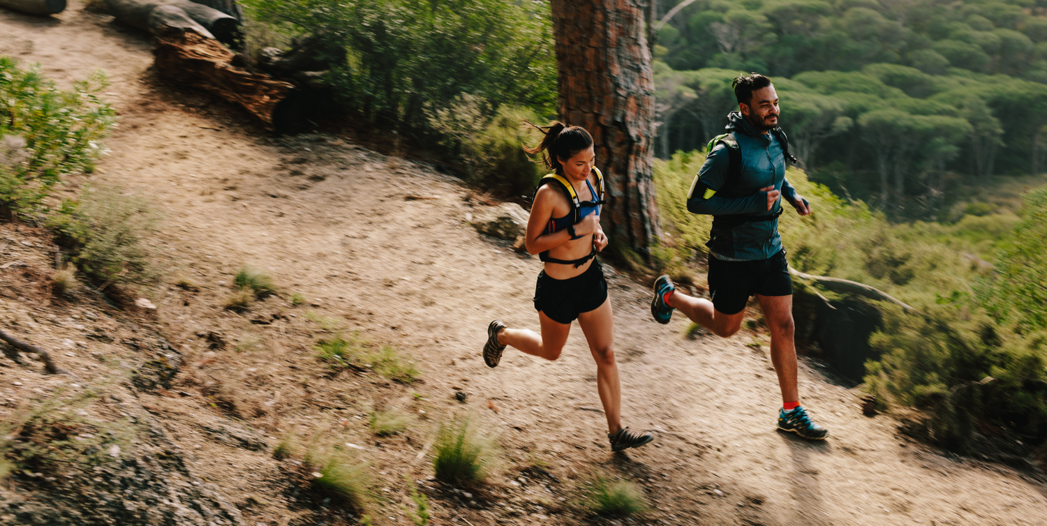 Male and female runners jogging on scenic outdoor trail surrounded by trees and nature.