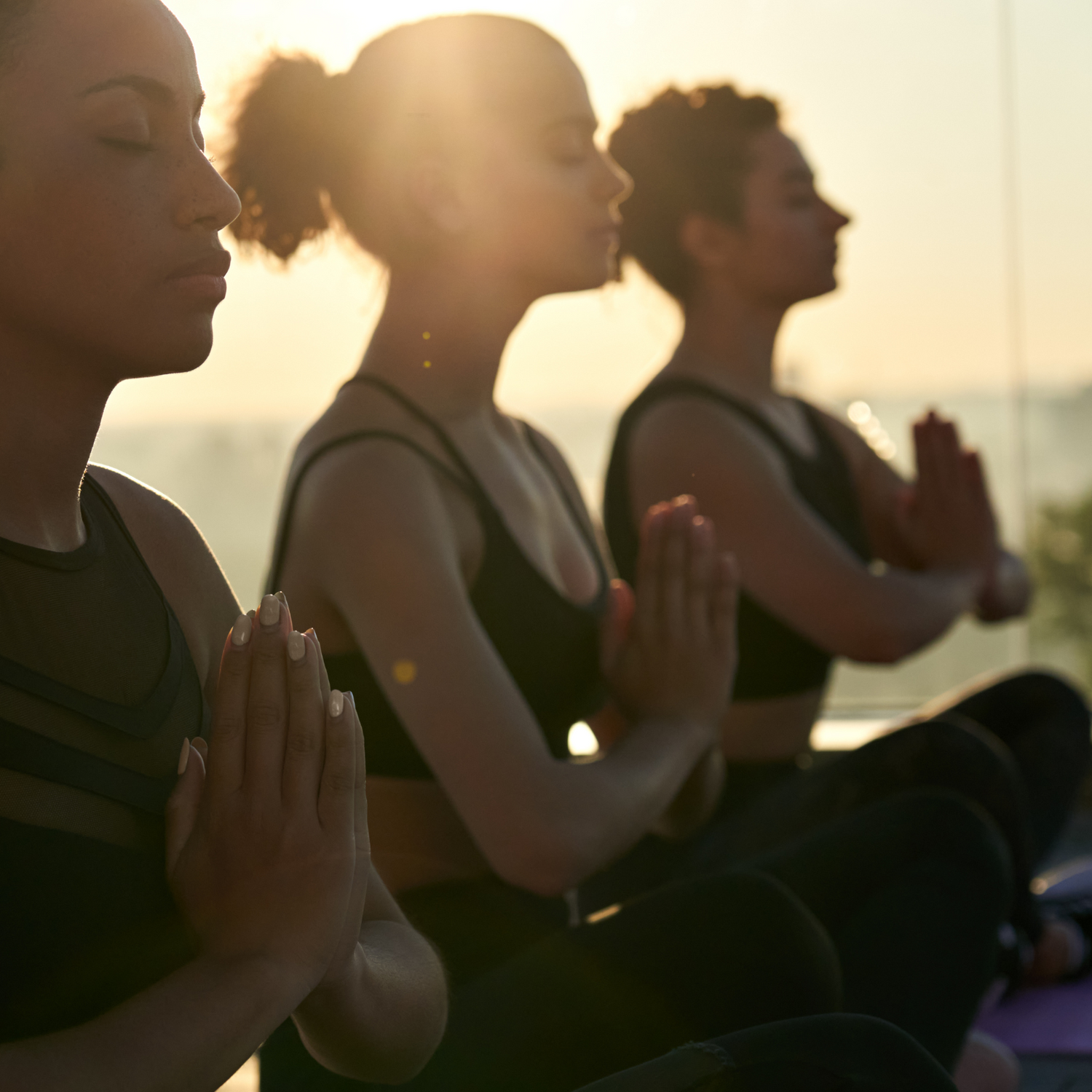 Three women sitting cross-legged in meditation pose, silhouetted against a golden sunrise with their eyes closed and hands together at their chests.