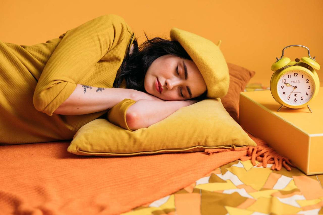Woman in soft pajamas sitting up in bed at night, lit by a warm bedside lamp, reading a book with a Soothe Organic Sleep gummy bottle on the nightstand.