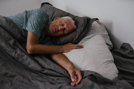 A smiling older woman in her 60s sleeping peacefully in a bright, comfortable bedroom, with a bottle of organic CBD gummies on her nightstand beside a glass of water — representing natural hemp-derived sleep support for seniors in Wyoming.