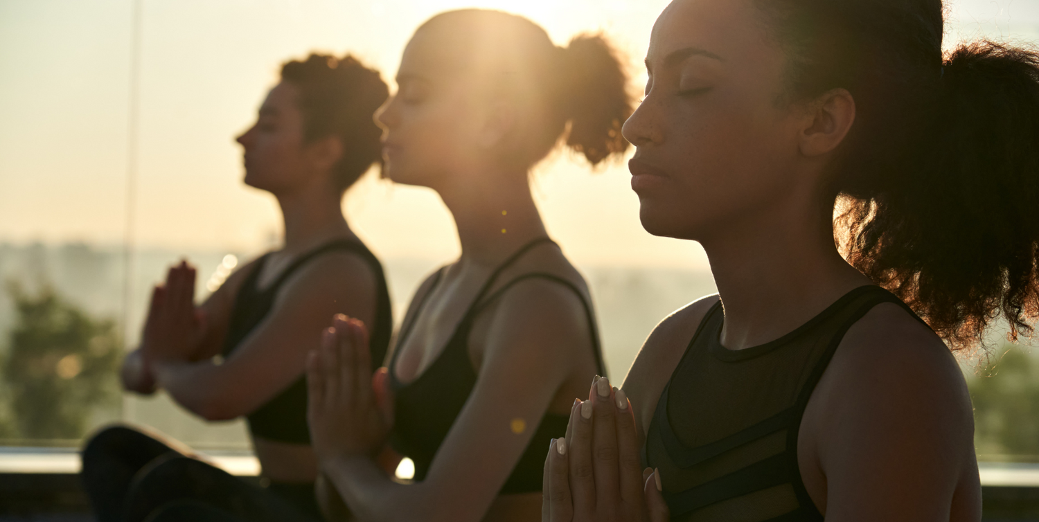 Three women sitting cross-legged in meditation pose, silhouetted against a golden sunrise with their eyes closed and hands together at their chests.."