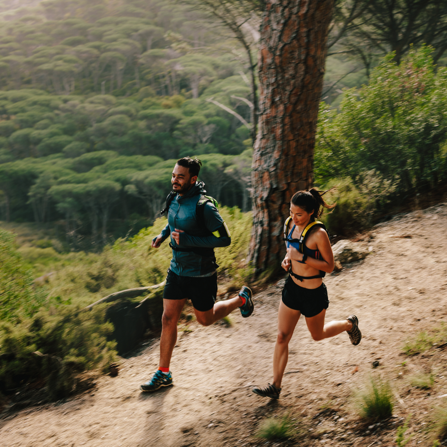 Male and female runners jogging on scenic outdoor trail surrounded by trees and nature.
